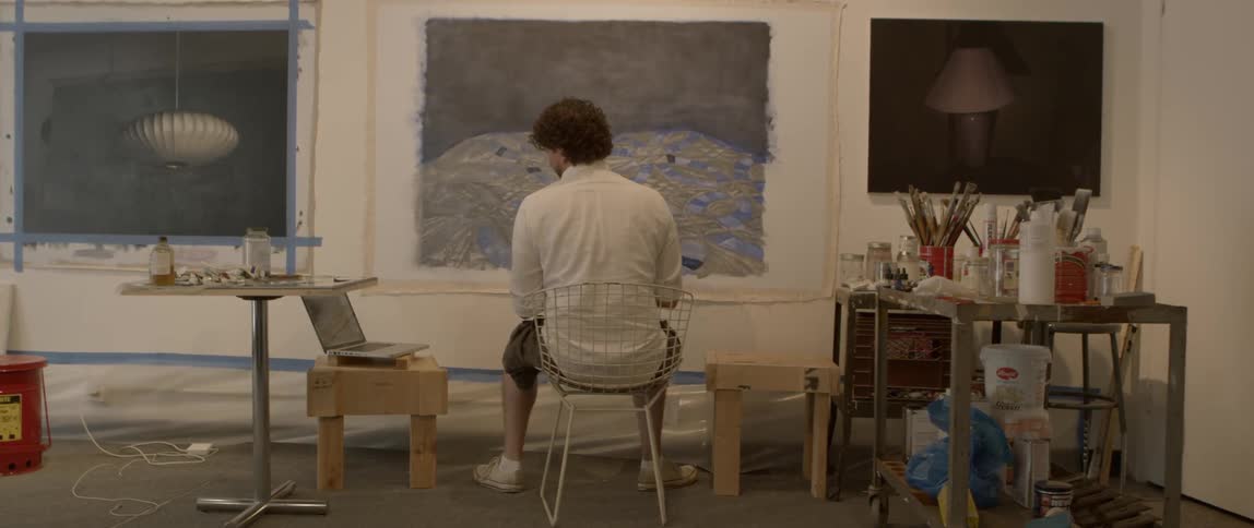 A man with curly brown hair sits in a small chair in front of a painting in progress on the wall in front of him before he reaches over with his brush to apply more paint to it
