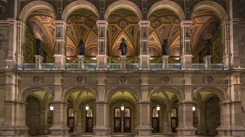 People walk by an old style building with statues inside of arches