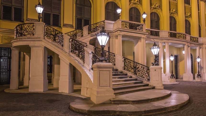 Lanterns light a grand staircase leading from a courtyard to a balcony level of a large yellow building at night