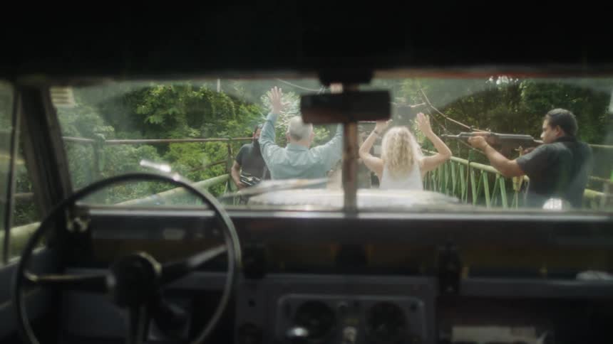 A man and woman walk along a bridge while men point guns at them