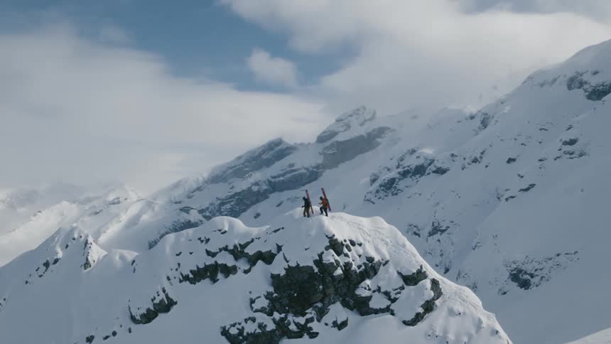 Two people stand a top a snow point with snowboards on their backs