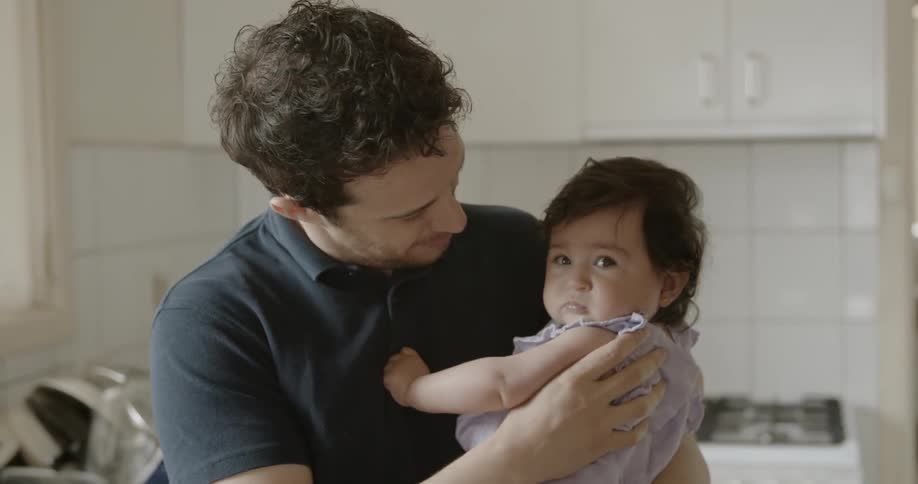 A father is holding a talking to his chubby infant daughter inside of a white kitchen