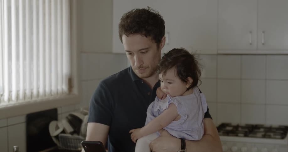 A man standing in a kitchen holding a baby girl while using a smartphone