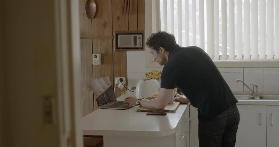 Man wearing black t shirt uses laptop on kitchen countertopduring daylight in the kitchen