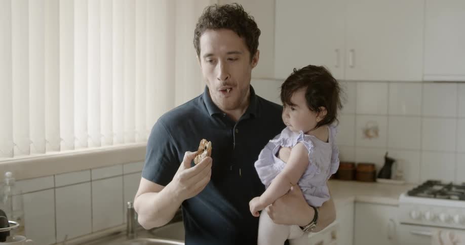 A father holds his daughter while eating a piece of bread in the kitchen