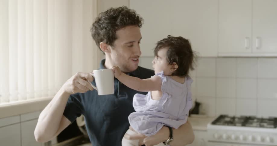 Man holding baby sips a cup of coffee in the kitchen during daylight