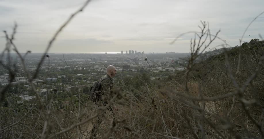 A white man with a beard hikes up a hill outside a city