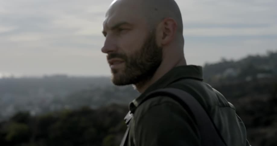 A white man with a beard stands and looks around at a city in a valley