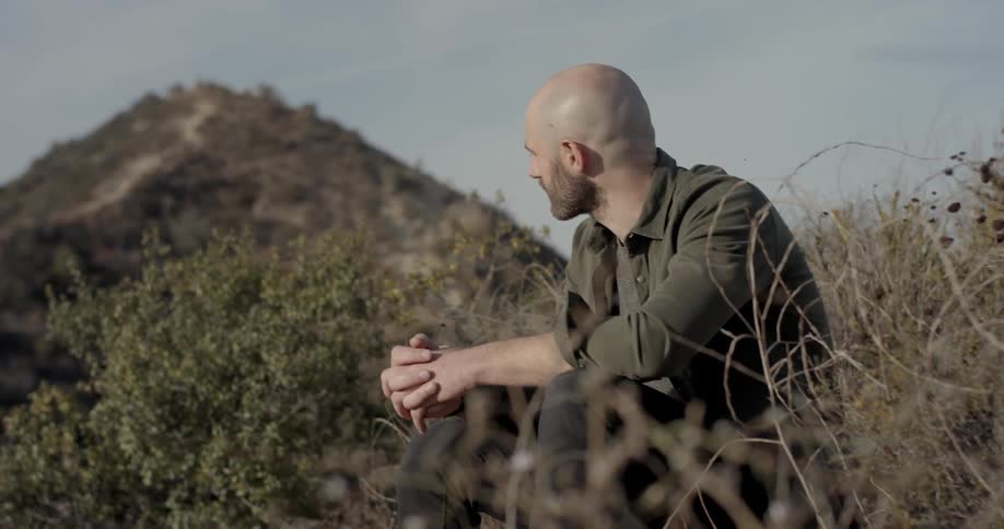 A white man with a beard sits on a hillside and looks at his surroundings