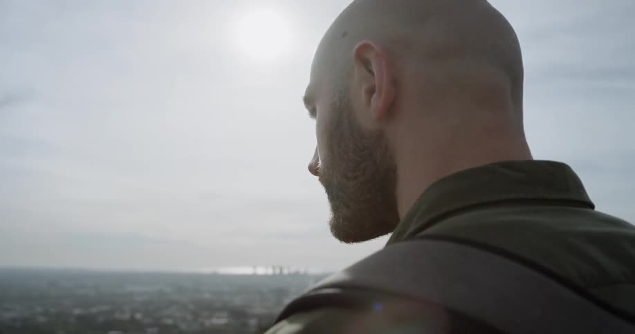 A bald man stands on a ridge and looks down at the city in a valley below on a sunny day