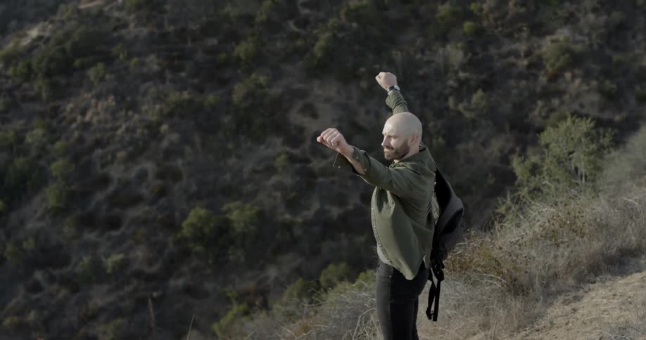 Bald bearded manwearing backpack walks on the mountain path and lifts his fists up during daylight