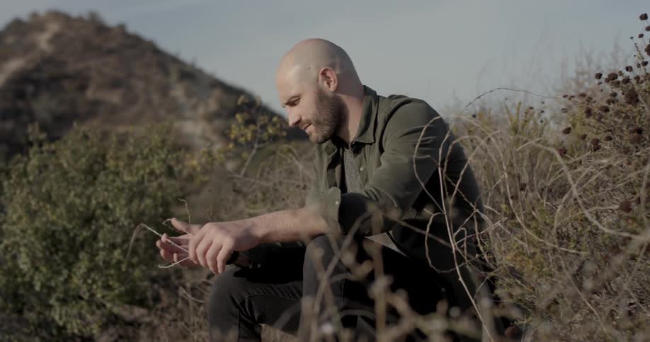 Man sits on hill among brush with mountain in background