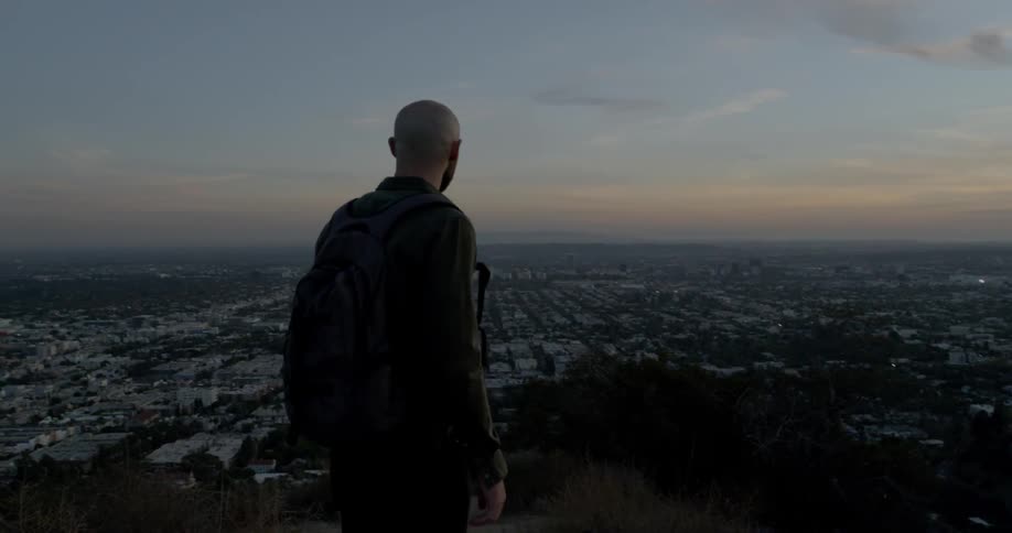 A hiker stands on a peak overlooking a dense city neighborhood at dusk
