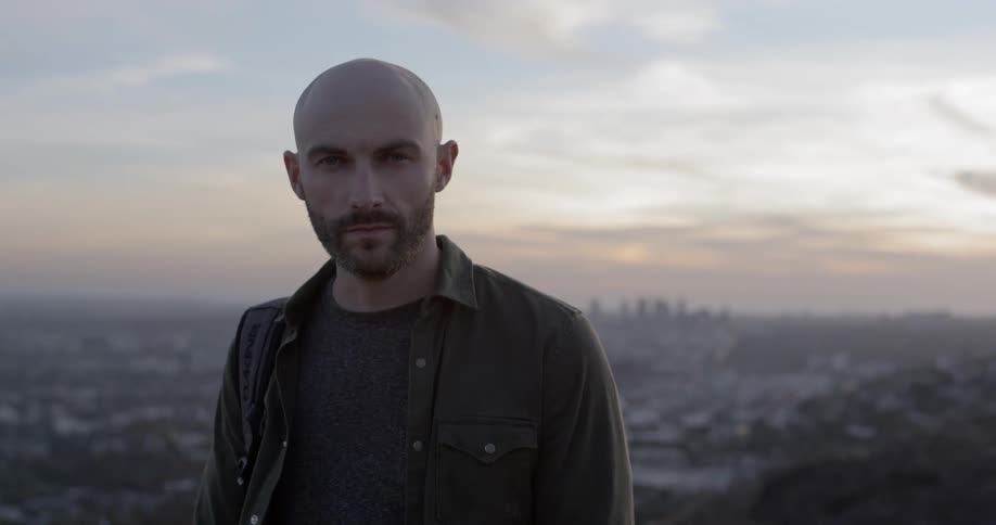 A white man looks into the camera from a high lookout point with a view of a city behind him
