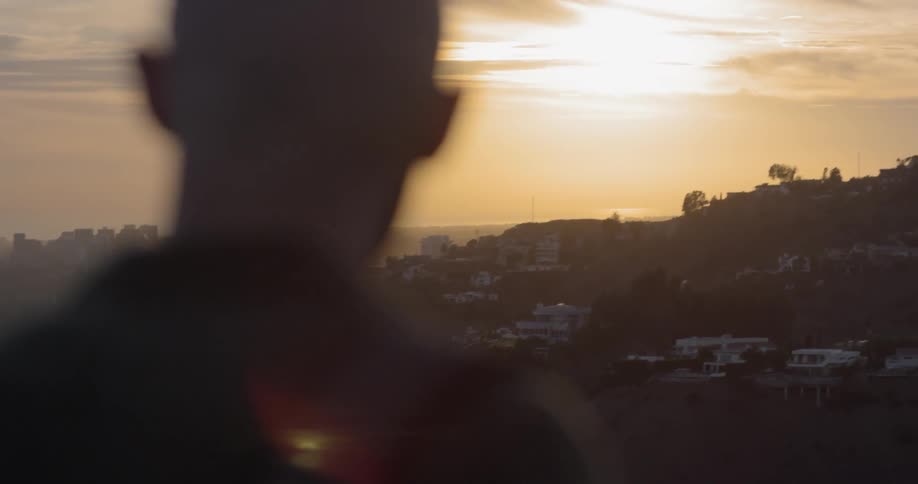 A man stands at the top of a mountain looking out at a mountain town in the distance at dusk