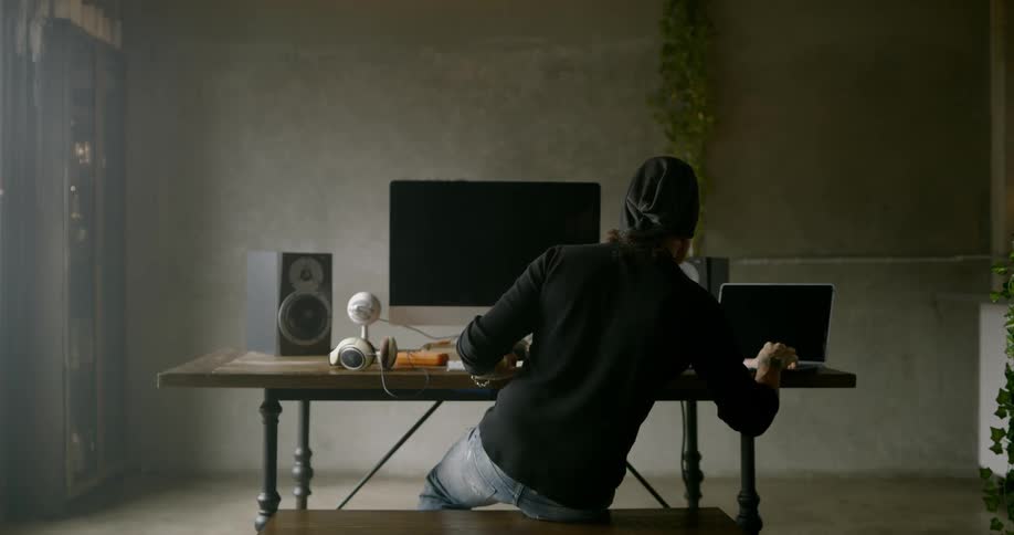 Man wearing black beanie and jeans sits at the table and uses laptop in a room during daylight