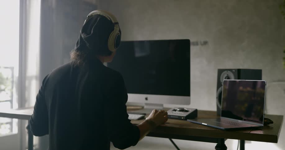Man sitting at the table in front of apple computer display wears headphones and shakes his head in a room during daylight