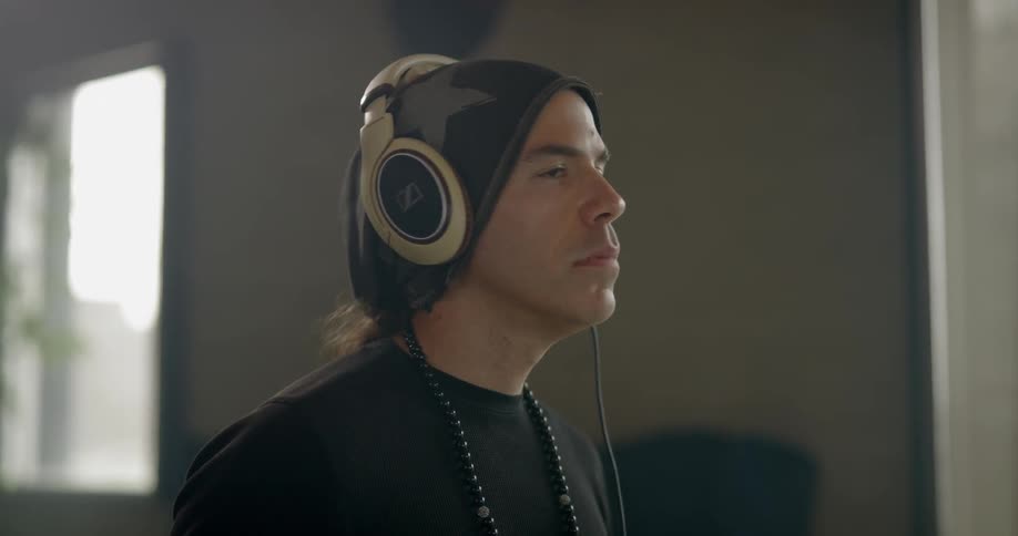 Man wearing black beanie and necklace listens to music through headphones indoors during daylight