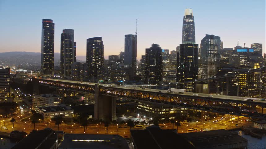Traffic streams across the upper and lower levels of a a suspension bridge that leads to a dense downtown area filled with many skyscrapers
