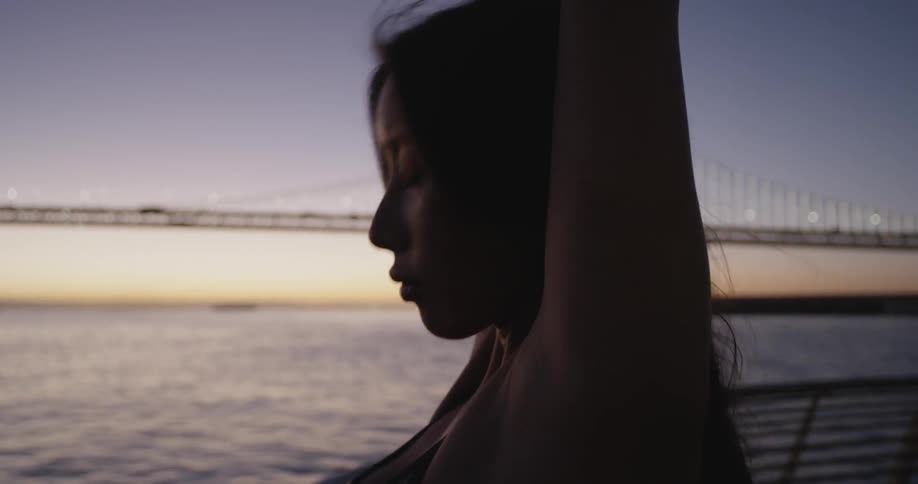 Woman stretches her arms on the promenade with the view of bridge at dawn
