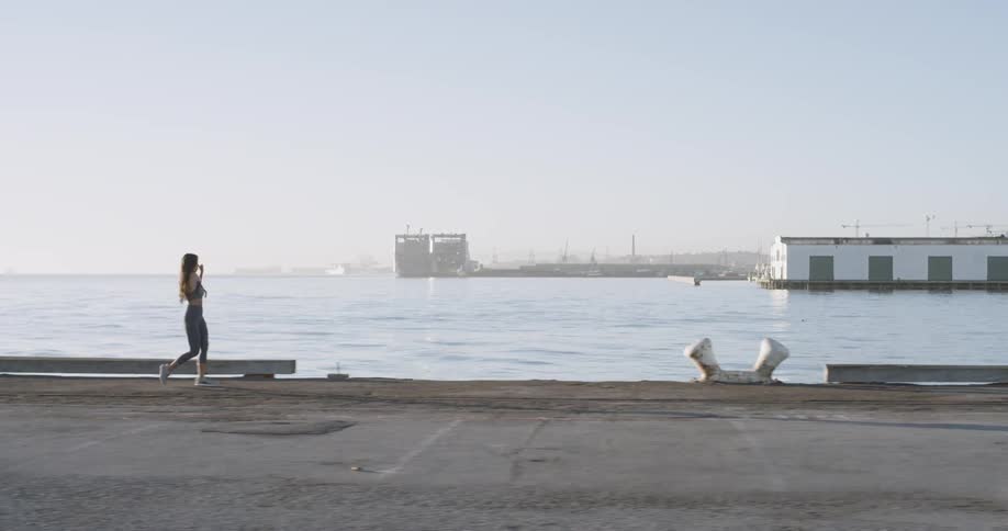 A female with long dark hair is stretching her shoulders and swinging her arms while walking along a pier next to the water