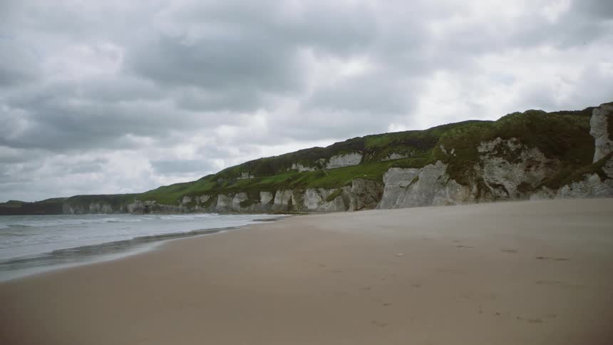 Small waves of the ocean hit the sandy beach next to the green hill on a cloudy day