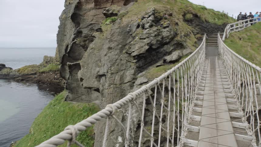 There is a small tour group at the end of the rope and wooden bridge that is suspended over the cliff and the ocean
