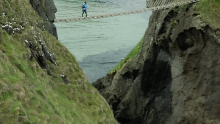 Now we see the height of the rope suspended bridge as we get a wide shot of the woman walking across it carefully while her husband waits on the other side and the seagulls fly