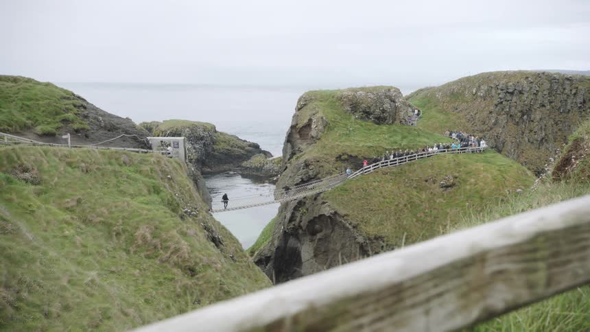 People waiting to cross rope bridge over water near ocean