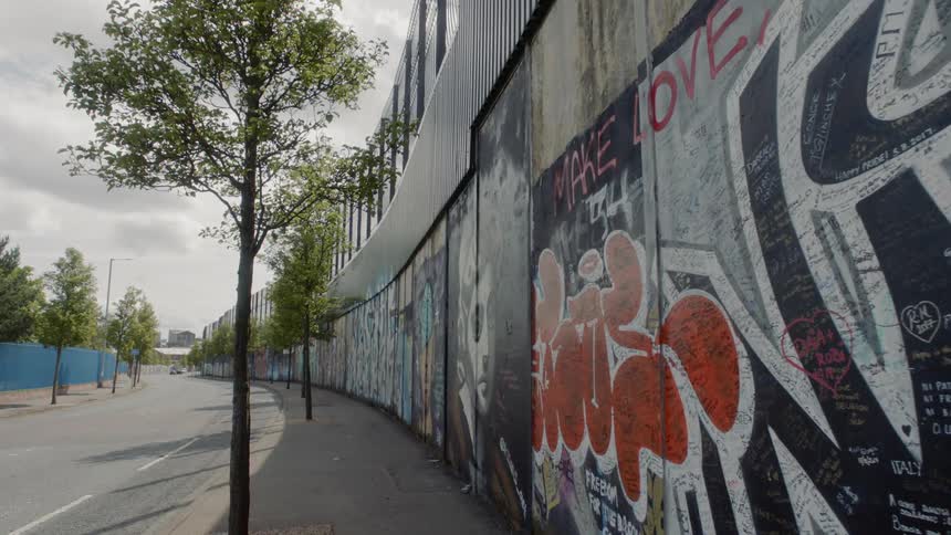 A wall with a fence on top is painted with graffiti and lines a street with young trees