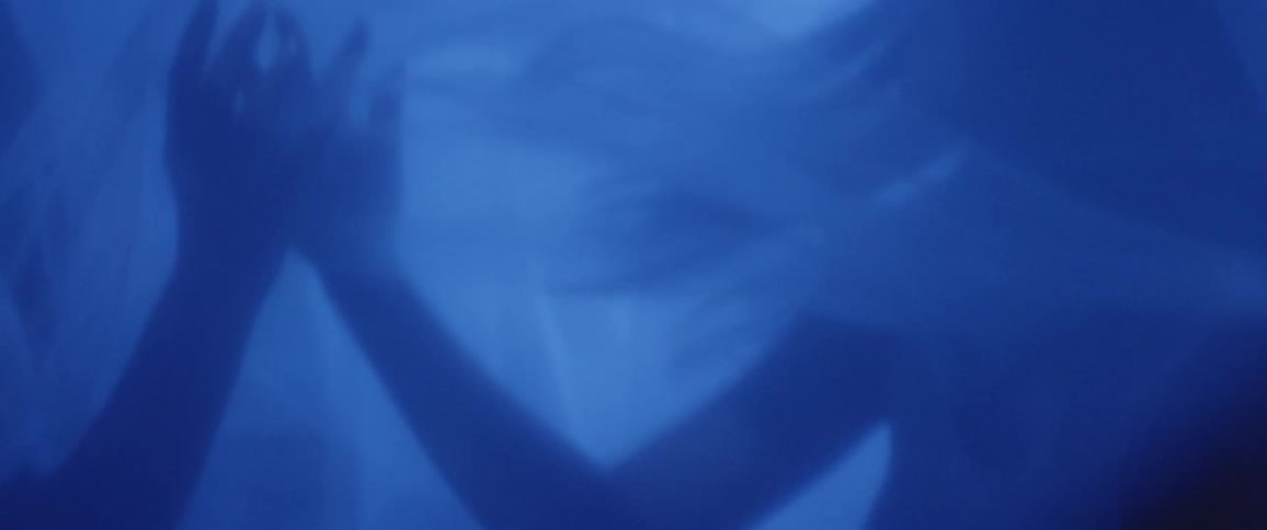 Two women dance and embrace in a tent with veils illuminated by electric blue light