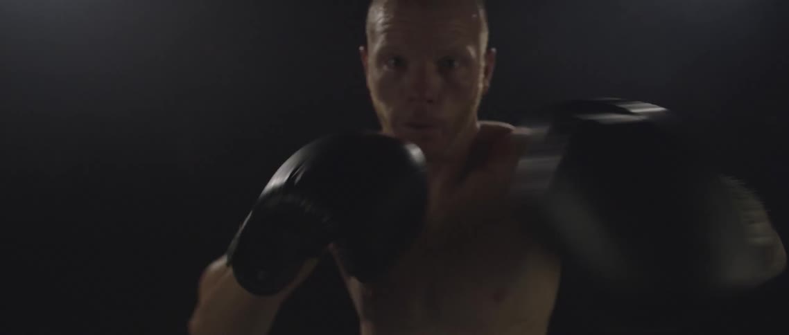 A shirtless athletic male in front of a black background punching towards the camera while wearing black boxing gloves