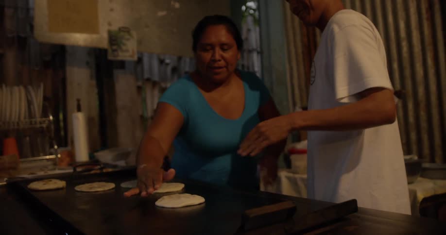 A man and a woman smiling while they cook flatbread on a large griddle
