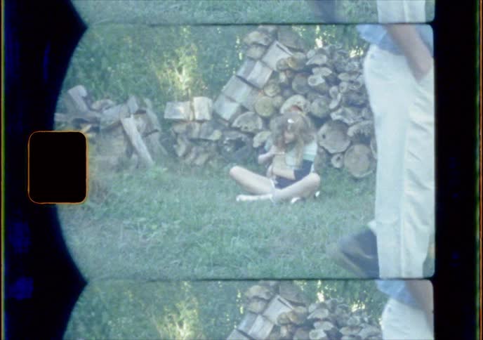 A girl sits in front of a woodpile petting a small animal while a man walks by