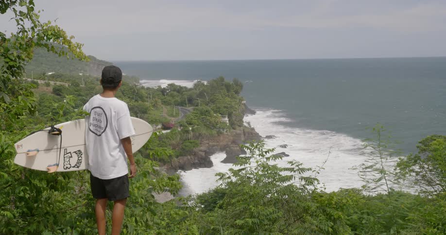 A person holds a surf board and looks down at crashing waves