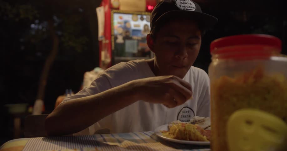 Asian boy sitting at the table eats food and finds it tasty outdoors at night