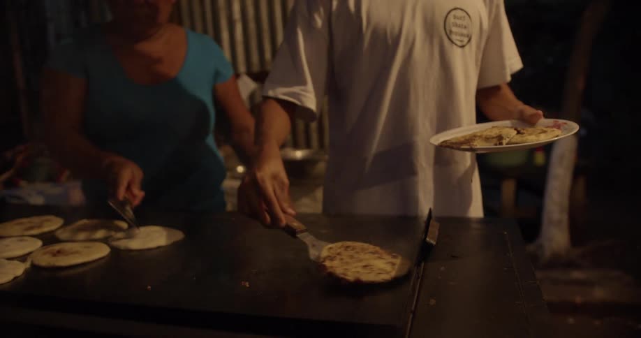A man takes finished pancakes onto onto a plate to serve them while a woman flips those pancakes still cooking
