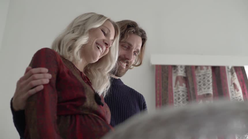 A man has his arm around a pregnant woman while they smile and look down at something in front of them