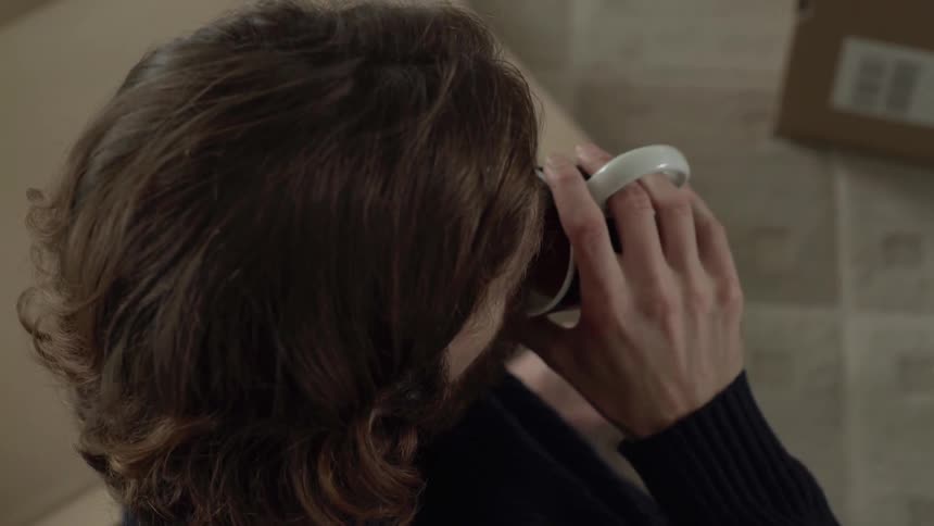 A white man in a sweater is drinking a cup of coffee and talking to someone