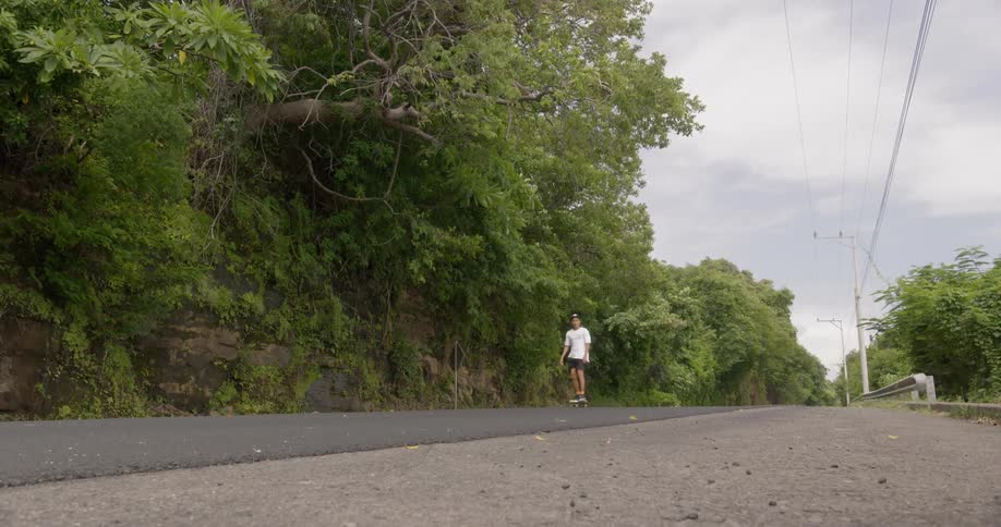 A man rides on a skateboard down an empty road that has a dense wall of trees and rocks on one side