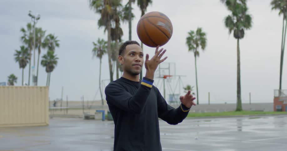 A man twirls a basketball on his finger while standing on a wet outdoor court with palm trees in the background