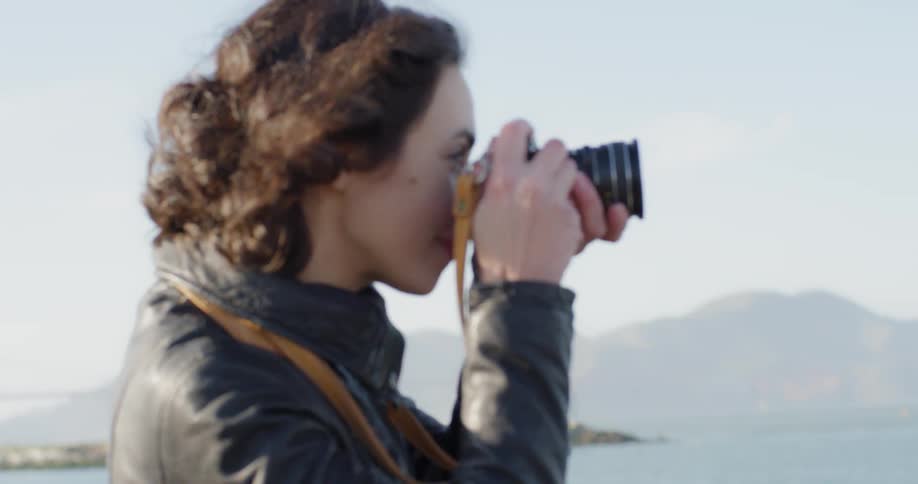 A white female in a leather jacket looks out over a body of water and takes a picture with a camera worn around her neck