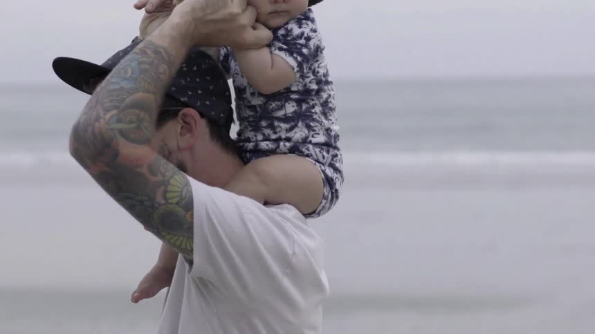 A man has a baby on his shoulders as he walks by the water on a cloudy day
