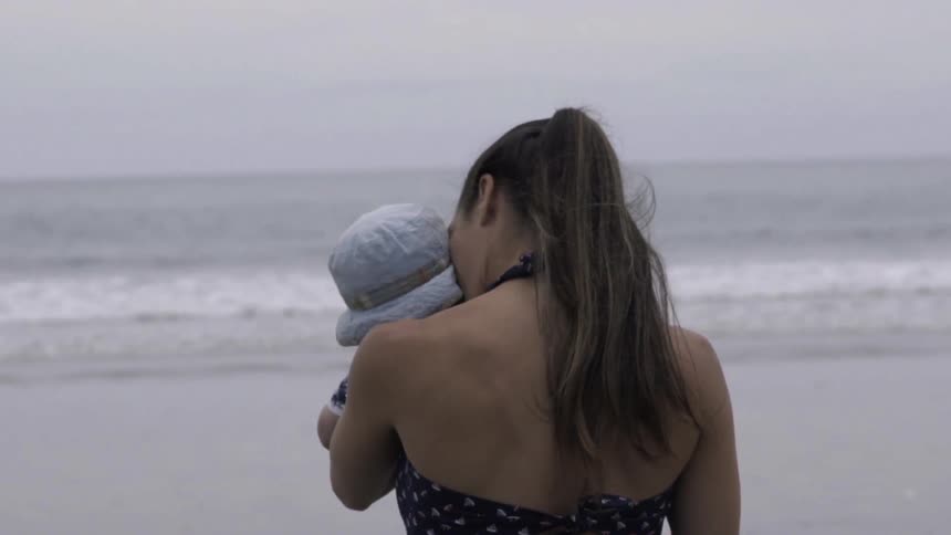 A woman kisses is a baby as she walks towards an ocean on a cloudy day