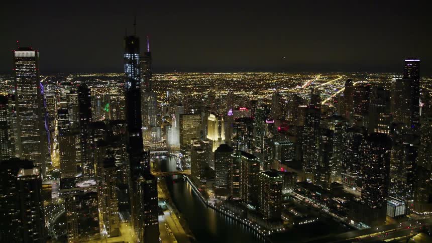A city skyline shimmers and glows at night as cars travel over the river