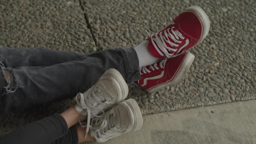 The legs and feet of a man and woman stretched out on the sidewalk with legs crossed and wearing canvas sneakers