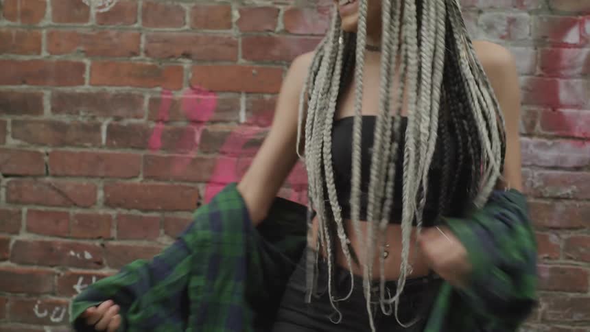 A young female with long dreadlocks dances in front of a brick wall with graffiti on it
