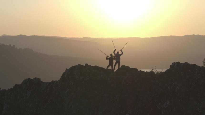 Two hikers hold their hiking sticks in the air while standing on a mountain ridge at sunrise