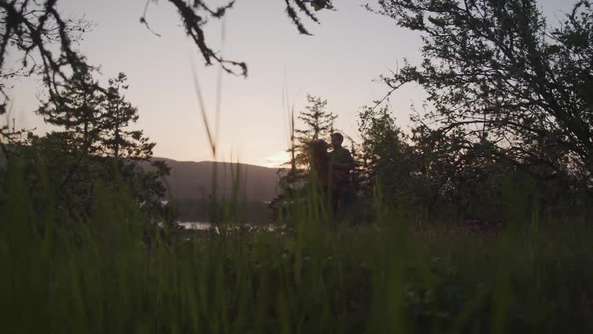 A man and woman stand up from the grass in an embrace at sunset