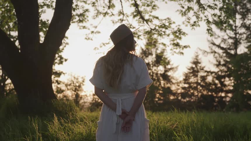 Woman wearing a sun hat stands in a grassy field near the trees as she looks around
